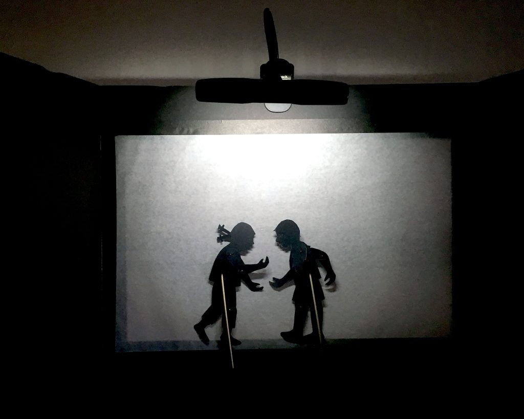 Inside of a shadow puppet screen showing a girl and boy puppet with holding rods 