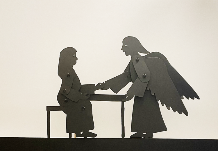 Silhouette puppets of Mary sitting at a table and an angel on the other side, holding her hand.
