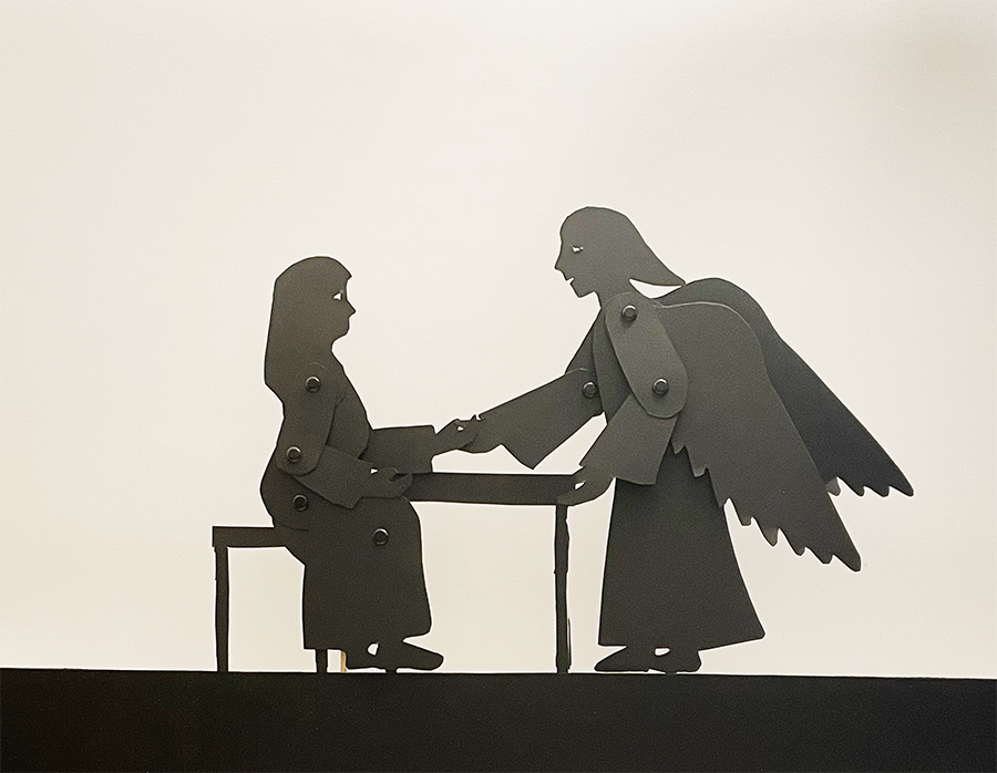 Silhouette puppets of Mary sitting at a table and an angel on the other side, holding her hand.